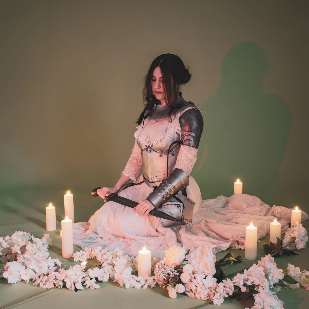 Deerest wearing armour pieces over a white dress, holding a sword across her lap, surrounded by candles and flowers.